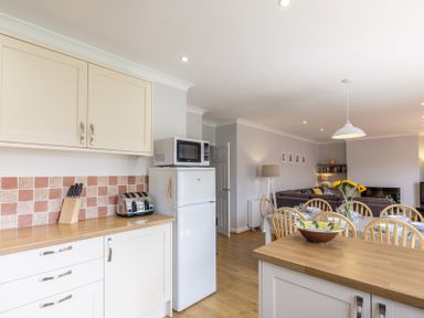 A kitchen with white cabinets a wooden countertop a refrigerator with a microwave on top and a dining table with chairs and flowers at Cogenhoe in Rock