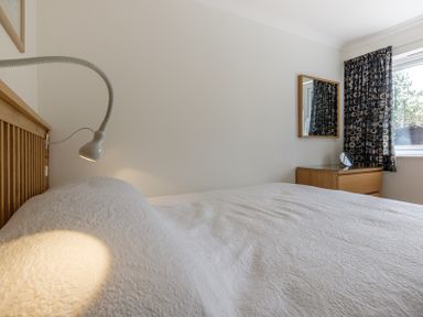 A bedroom with a bed covered by a white bedspread a wooden headboard a bedside lamp a wooden dresser a mirror and a window with patterned curtains at Cogenhoe in Rock