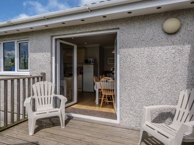 A wooden deck with two plastic chairs outside a building with open double doors showing a dining table and kitchen inside at Cogenhoe in Rock