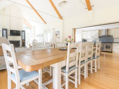 A dining room with a wooden table and white chairs next to an open kitchen with white cabinets and stainless steel stove at Ragleighs in Daymer Bay