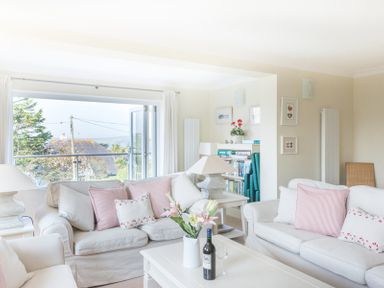 A living room with white sofas pink cushions flowers on a table and a view outside at Ragleighs in Daymer Bay