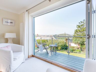 A room with a white armchair and lamp opening onto a balcony with a chair and small table at Ragleighs in Daymer Bay