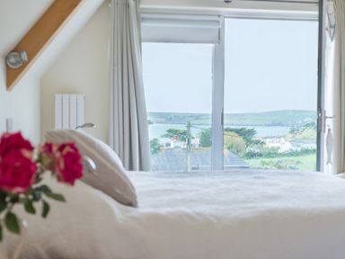 A bedroom with a bed white bedding and a window with curtains showing a view of houses trees and a body of water at Ragleighs Daymer Bay