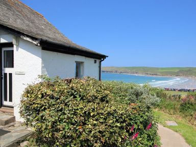 Exterior view of a white cottage with bushes in front and a beach with waves and green hills in the background at Gullsway Kittiwake in Polzeath