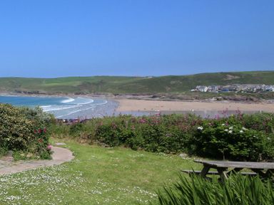 A grassy area with bushes and a wooden picnic table overlooking a sandy beach and ocean with hills and buildings in the background at Gullsway September Tide in Polzeath