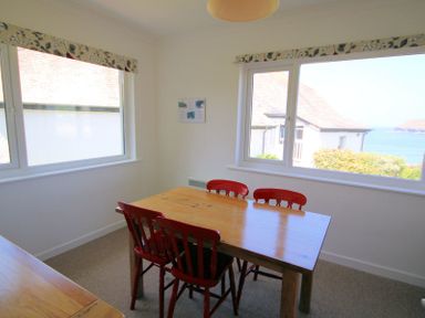 A dining room with a wooden table and red chairs by windows overlooking houses and the sea at Gullsway September Tide Polzeath