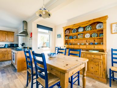 A kitchen dining area with a wooden table and blue chairs wooden cupboards and shelves with decorative plates at Little Sailing in Rock