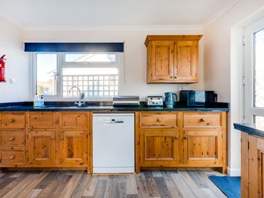 A kitchen with wooden cabinets a white dishwasher a toaster a kettle a microwave and a window with a blue blind at Little Sailing in Rock