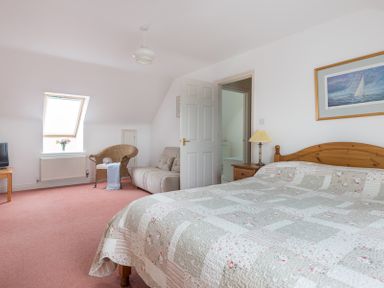 A bedroom with a wooden bed a side table with a lamp a sofa a wicker chair a small table a television and a window at Kirra in Polzeath