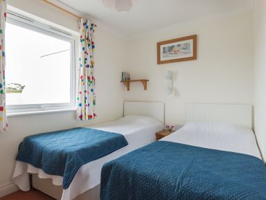 A bedroom with two single beds with blue quilts a window with polka dot curtains a small wooden shelf and a framed picture on the wall at Kirra in Polzeath