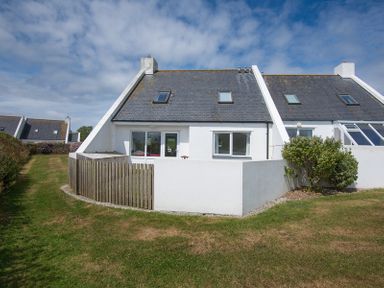 A white house with a grey sloped roof and small fenced patio in front with grass surrounding the property at Kirra in Polzeath