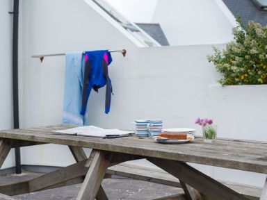 An outdoor wooden picnic table with mugs plates a map and a wetsuit hanging on a rail at Kirra in Polzeath