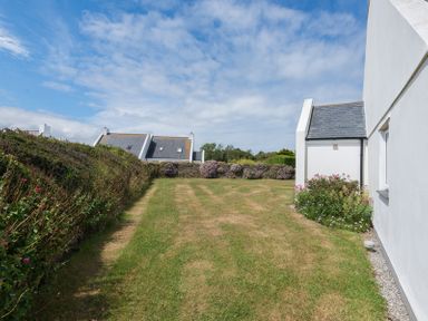 A garden with a lawn bordered by hedges and flowering plants next to a white building at Kirra in Polzeath