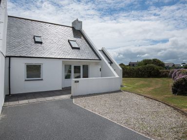 A white house with a slate roof driveway and gravel area next to a lawn with purple flowers at Kirra in Polzeath