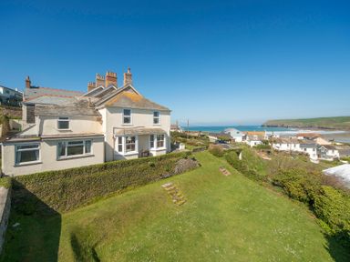 A white house with chimneys on a hill overlooking a grassy yard and coastal village at Pentire View 3 in Polzeath