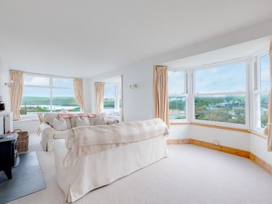 A living room with cream sofas, large windows with curtains, and views of hills and houses at Pentire View (3) in Polzeath