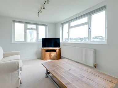 A living room with a white sofa a wooden coffee table a corner wooden TV stand with a television and large windows at Pentire View (3) in Polzeath