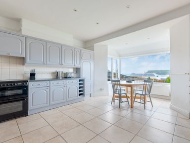 A kitchen with blue cabinets a black stove a wooden dining table with three chairs and a large window showing an outdoor view at Pentire View (3) in Polzeath