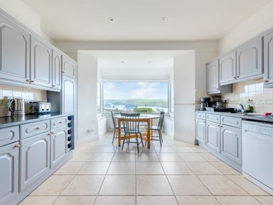 A kitchen with gray cabinets, a round wooden table with chairs, and a large window showing a view outside at Pentire View (3) in Polzeath