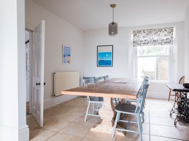 A dining room with a wooden table and six chairs two framed pictures on the walls and a window with patterned blind at Pentire View (3) in Polzeath