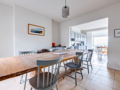 A dining area with a wooden table and gray chairs leading to a kitchen with gray cabinets at Pentire View (3) in Polzeath