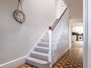 A hallway with a carpeted staircase and a wall clock overlooking a patterned tile floor leading to a kitchen at Pentire View (3) Polzeath