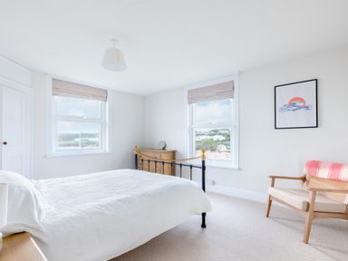 A bedroom with a bed chair dresser two windows and wall art at Pentire View (3) in Polzeath