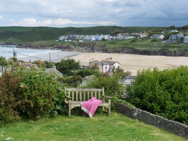A wooden bench with a pink blanket on a grassy hill overlooking a beach and houses at Pentire View (3) in Polzeath