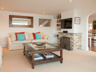 A living room with beige sofas red and blue cushions a wooden coffee table and a stone fireplace with a wall-mounted TV at Treverden in Polzeath