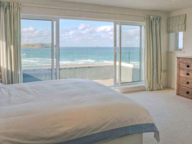 A bedroom with a bed facing sliding glass doors overlooking the ocean and a wooden dresser to the right at Treverden in Polzeath
