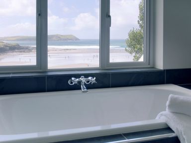 A bathtub with towels beside it and a window showing a beach and ocean view at Treverden in Polzeath