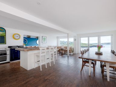 An open kitchen and dining area with wooden floors and a large table with chairs near windows overlooking a coastal landscape at Treverden Polzeath