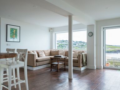 A living room with a beige corner sofa and wooden coffee table by a large window and glass door showing a coastal view at Treverden in Polzeath