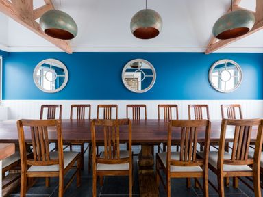 A dining room with a long wooden table and twelve wooden chairs under three hanging lights and three round mirrors on a blue wall at Treverden Polzeath