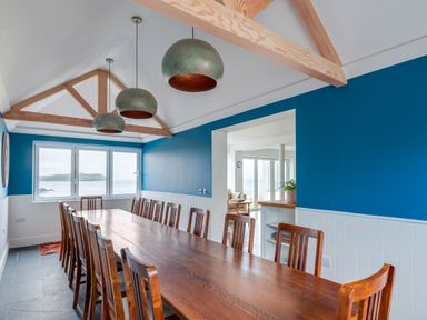 A dining room with a long wooden table and chairs under a vaulted ceiling with wooden beams and three hanging lights overlooking the sea at Treverden in Polzeath