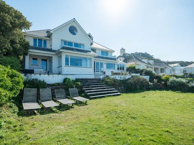 A white multi-story house with large windows and a circular window above stairs leading down to a lawn with five wooden lounge chairs at Treverden in Polzeath