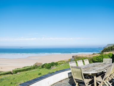 An outdoor patio with wooden table and chairs overlooking a sandy beach and ocean at Treverden in Polzeath