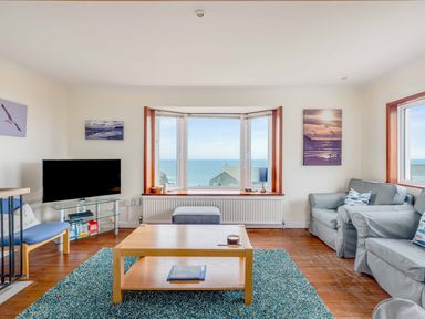 A living room with a wooden coffee table a blue rug two grey armchairs a blue chair a flat screen TV and a large window with sea view at Shilling Stones in Port Isaac