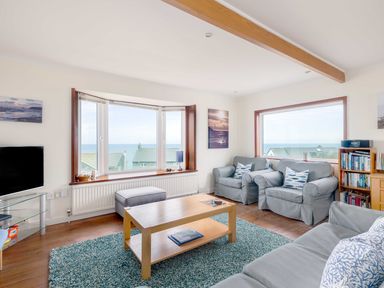 A living room with gray armchairs a wooden coffee table a blue rug and windows overlooking the sea at Shilling Stones in Port Isaac
