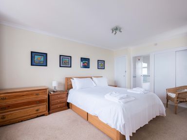 A bedroom with a double bed white linens wooden dressers framed pictures on the wall and a wicker chair at Shilling Stones in Port Isaac