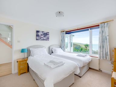 A bedroom with two single beds next to a window with curtains and a view of greenery and water at Shilling Stones in Port Isaac