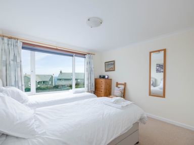 A bedroom with two single beds a wooden dresser a chair with cushion a wall mirror and a large window with curtains at Shilling Stones in Port Isaac