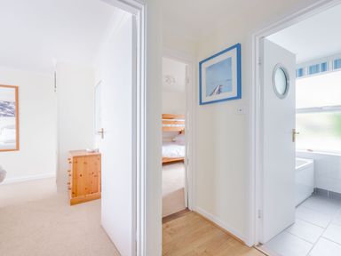 A hallway with three open doors showing a bedroom with a wooden dresser another bedroom with bunk beds and a bathroom with a bathtub at Shilling Stones in Port Isaac