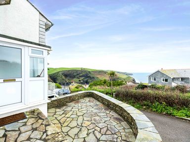 A stone patio with a low stone wall overlooking green hills and the sea at Shilling Stones in Port Isaac