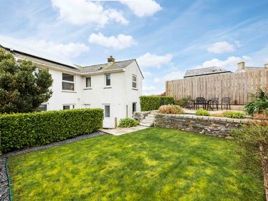 A backyard with grass lawn a white two story house stone retaining wall and outdoor seating area at Shilling Stones in Port Isaac