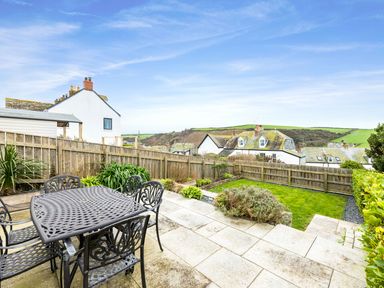 A patio with a metal table and chairs overlooking a fenced garden with bushes and grass at Shilling Stones in Port Isaac