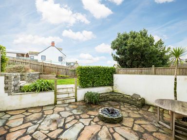 An outdoor patio area with stone flooring steps leading to a grassy garden plants a wooden table and a stone fire pit at Shilling Stones in Port Isaac