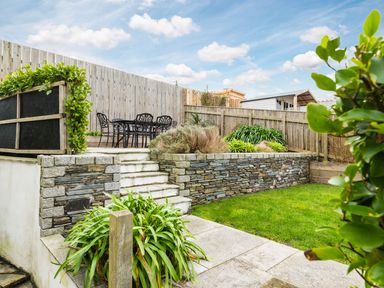 A garden with a stone retaining wall wooden fence a patio with a table and chairs and green plants at Shilling Stones in Port Isaac