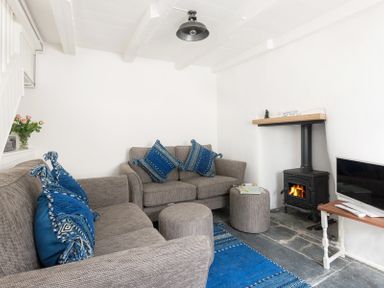 A living room with two gray sofas with blue cushions a wood stove a small wooden table with a television and a blue rug at Bre Cottage in Port Isaac