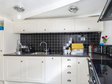 A kitchen with white cabinets black tiled backsplash a sink and faucets a kettle bread bin knives microwave and flowers on the counter at Bre Cottage in Port Isaac